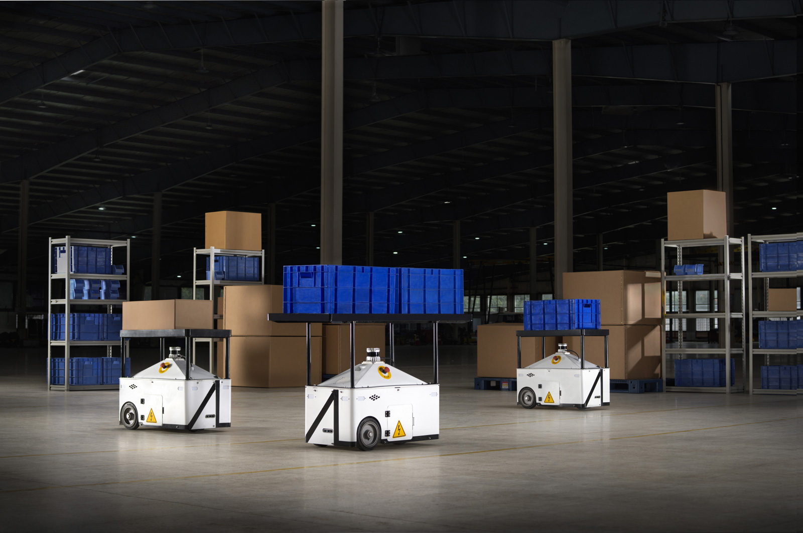 Three white autonomous robots carry blue storage bins inside a spacious warehouse with shelves stacked with brown and blue boxes in the background. The area is well-lit and organized.