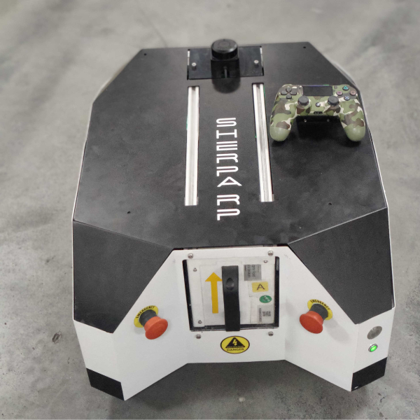 A black and white robotic device with a game controller on top, featuring control buttons, warning labels, and geometric symbols. The device rests on a gray concrete floor.