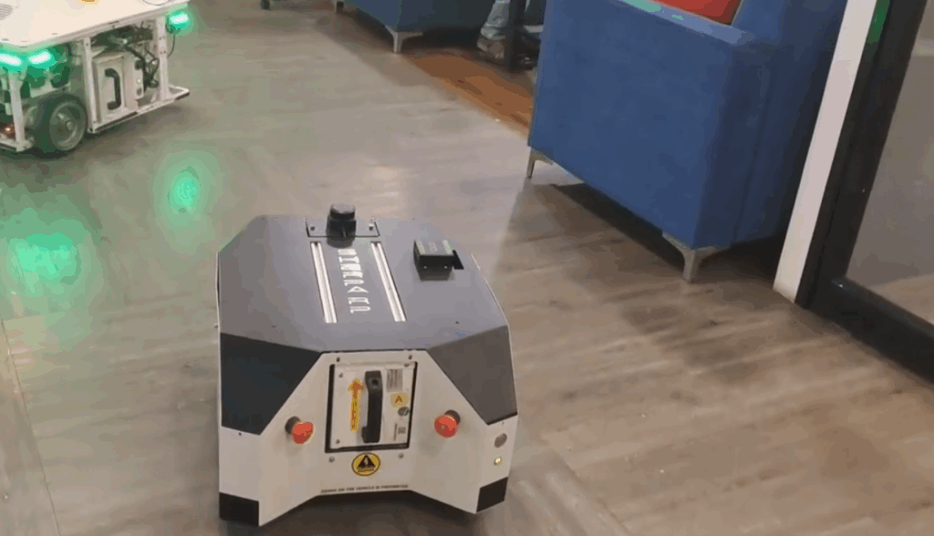 A small, boxy mobile robot with black and white panels and warning labels moves across a room with wooden floors, near blue chairs and another robot in the background.