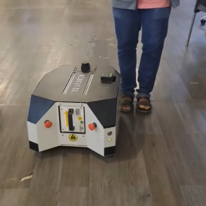 A person in jeans and sandals stands behind a small, boxy robotic device with buttons and controls on its surface, positioned on a wooden floor in an indoor setting.